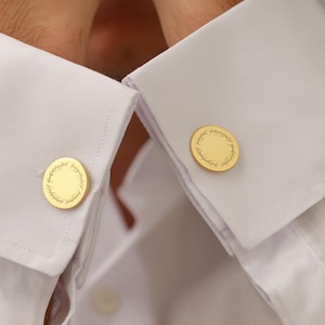May include: Gold-colored cufflinks with a circular design and text, fastened to the cuffs of a white dress shirt. The cufflinks have a detailed, engraved pattern around the edge.