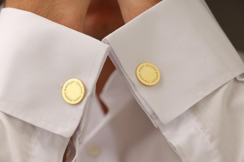 May include: Gold-colored cufflinks with a circular design and text, fastened to the cuffs of a white dress shirt. The cufflinks have a detailed, engraved pattern around the edge.