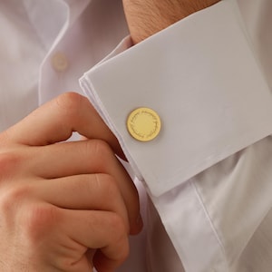 May include: Gold-colored cufflinks with a circular design are fastened on a white dress shirt. The cufflinks have a detailed, textured pattern. The shirt's cuff is folded over, and the person's hand is visible.