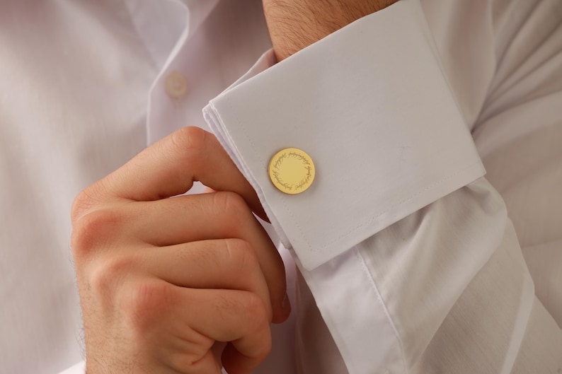 May include: Gold-colored cufflinks with a circular design are fastened on a white dress shirt. The cufflinks have a detailed, textured pattern. The shirt's cuff is folded over, and the person's hand is visible.