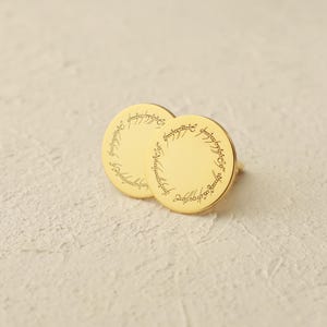 May include: Gold-colored round stud earrings. Each earring features an engraved inscription around the edge. The earrings are displayed on a textured, off-white surface. The design is reminiscent of a fantasy theme.