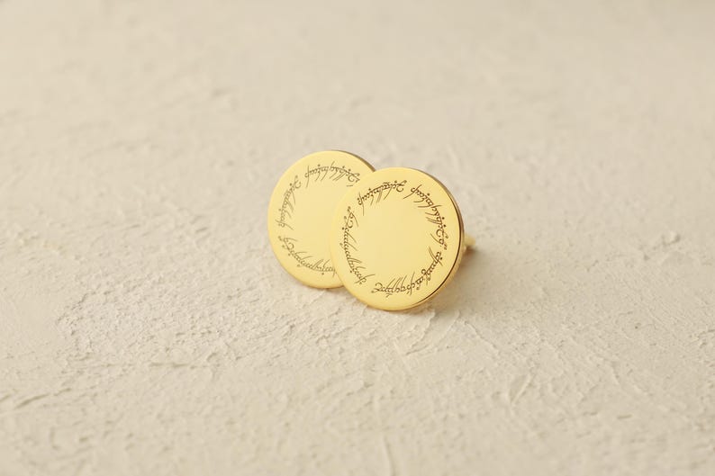 May include: Gold-colored round stud earrings. Each earring features an engraved inscription around the edge. The earrings are displayed on a textured, off-white surface. The design is reminiscent of a fantasy theme.
