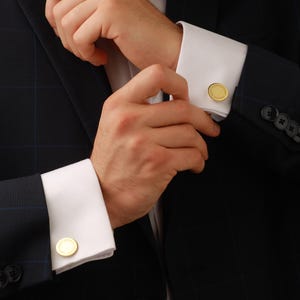 May include: Close-up of a dark navy blue suit jacket with a subtle plaid pattern. The white shirt cuffs are adorned with gold-colored, round cufflinks. The jacket has black buttons and a classic, formal style.
