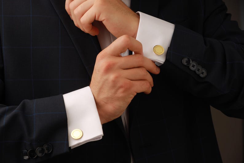 May include: Close-up of a dark navy blue suit jacket with a subtle plaid pattern. The white shirt cuffs are adorned with gold-colored, round cufflinks. The jacket has black buttons and a classic, formal style.