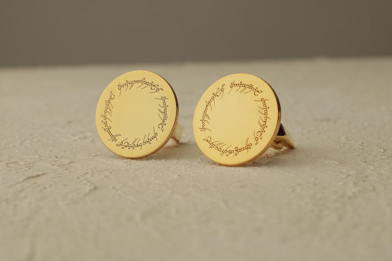 May include: Two gold-colored rings with a circular design and engraved text around the edge. The rings are set against a neutral background. The design and text are reminiscent of a fantasy theme.