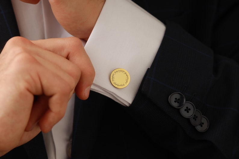 May include: Gold-colored cufflinks with a circular design and engraved text. The cufflinks are on a white dress shirt cuff, paired with a dark blue suit jacket. The image shows a close-up of the cuff and the person's hand.
