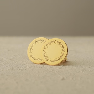May include: A pair of gold-colored round earrings with engraved text around the edges. The earrings are displayed on a textured, off-white surface. The text appears to be in a script or stylized font.