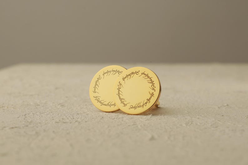 May include: A pair of gold-colored round earrings with engraved text around the edges. The earrings are displayed on a textured, off-white surface. The text appears to be in a script or stylized font.