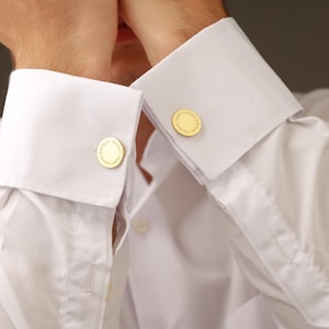 May include: Close-up of a white dress shirt with gold cufflinks. The cufflinks are round with a detailed design. The shirt has a classic collar and buttoned cuffs, suggesting formal wear.
