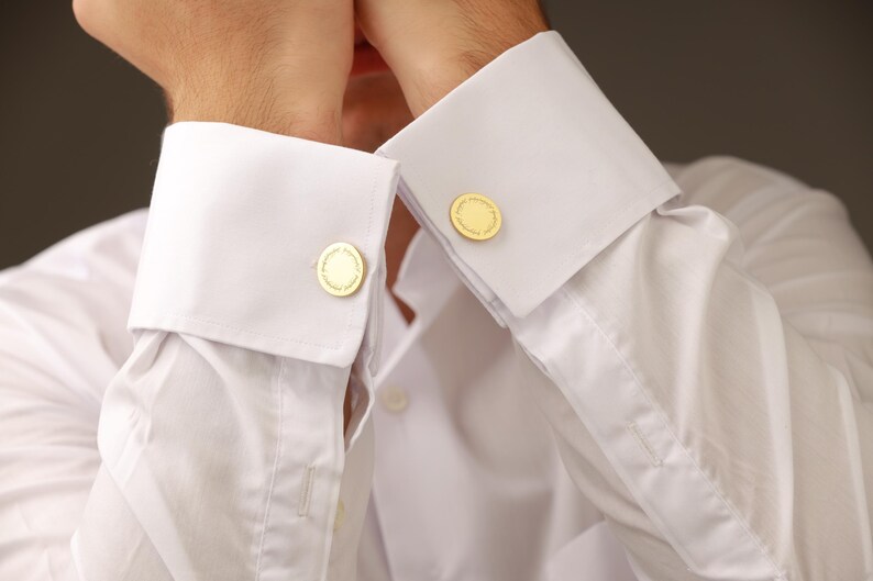 May include: Close-up of a white dress shirt with gold cufflinks. The cufflinks are round with a detailed design. The shirt has a classic collar and buttoned cuffs, suggesting formal wear.