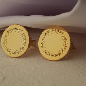 May include: A pair of gold-colored cufflinks with a circular design. Each cufflink features an engraved inscription around the edge. The cufflinks are resting on a textured surface, with a white fabric in the background.