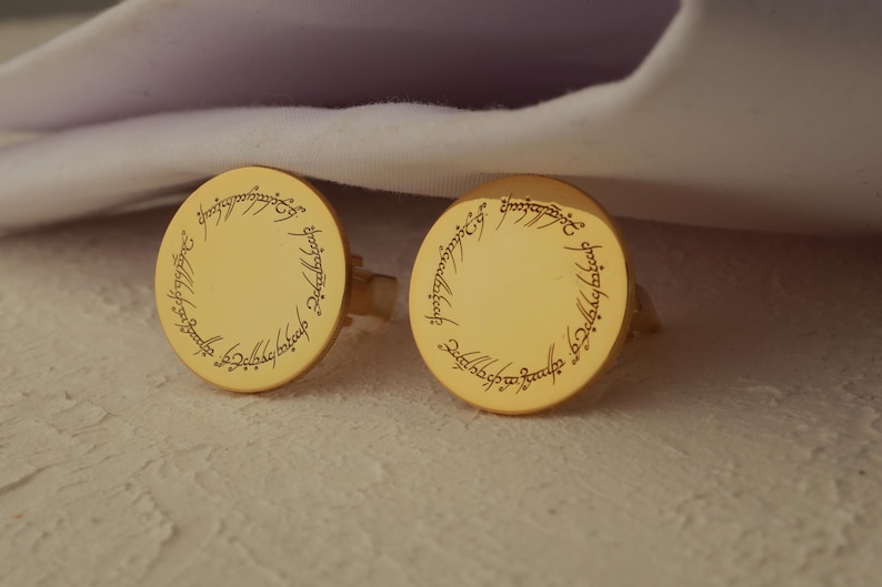 May include: A pair of gold-colored cufflinks with a circular design. Each cufflink features an engraved inscription around the edge. The cufflinks are resting on a textured surface, with a white fabric in the background.