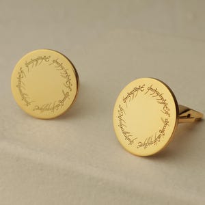 May include: A pair of gold-colored cufflinks. Each cufflink is round with a polished surface and features an engraved inscription around the edge. The cufflinks are displayed on a light-colored surface.