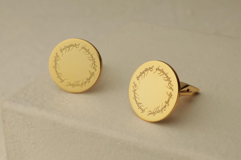 May include: A pair of gold-colored cufflinks. Each cufflink is round with a polished surface and features an engraved inscription around the edge. The cufflinks are displayed on a light-colored surface.