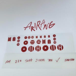 May include: A sheet of red vinyl decals featuring the word Arirang in a stylised script font, various geometric circular and rectangular symbols, and the names RM, JIN, SUGA, J-HOPE, JIMIN, and JUNG KOOK written in a handwritten red font.