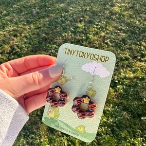 Könnte beinhalten: Ein Paar Donut-Ohrringe mit Schokoladenfrosting, Streuseln und einem gelben Stern auf einer weißen Karte mit dem Text "TINYTOKYOSHOP" und "HAND CRAFTED IN CANADA".