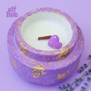 Peut inclure: Une bougie de couleur lavande dans un récipient rond avec des accents de feuilles d'or. La bougie a un intérieur en cire blanche, un bâton de cannelle et une décoration en forme de cœur violet. Le texte "elf hub" est inclus.