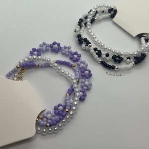 Op de afbeelding: Twee kralenarmbanden, een met paarse en witte kralen en de andere met zwarte en witte kralen. Beide armbanden hebben een bloemenpatroon.