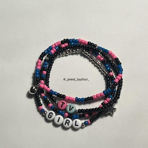 May include: A set of beaded bracelets with black, pink and blue beads. The bracelets spell out "TV GIRL" with white letter beads. There is a silver star charm and a silver heart charm on the bracelets.