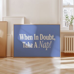 May include: A framed print with a blue background and the text "When In Doubt, Take A Nap!" in a serif font. The frame is a light wood colour. The print is displayed in a room with a window and hardwood flooring.