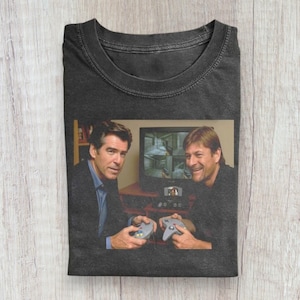 Peut inclure: T-shirt gris foncé avec une photo de deux hommes jouant à un jeu vidéo. L'image montre les hommes tenant des manettes et regardant un écran de télévision. Le t-shirt est fait d'une matière douce et a un col rond classique.