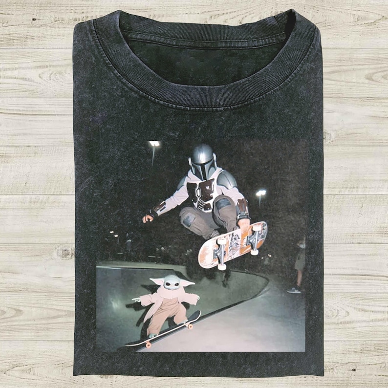 May include: Dark gray t-shirt featuring a graphic of The Mandalorian and Grogu skateboarding in a skate park. The image shows the characters mid-air and on a ramp. The shirt has a vintage, worn look.