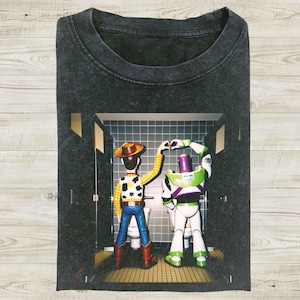K&ouml;nnte beinhalten: Dunkelgraues T-Shirt mit einer Grafik von Woody und Buzz Lightyear aus Toy Story. Die Charaktere befinden sich in einer Badezimmerumgebung, Woody tr&auml;gt einen Cowboyhut und Buzz seinen Raumanzug. Das Bild ist auf einem weichen, l&auml;ssigen T-Shirt gedruckt.