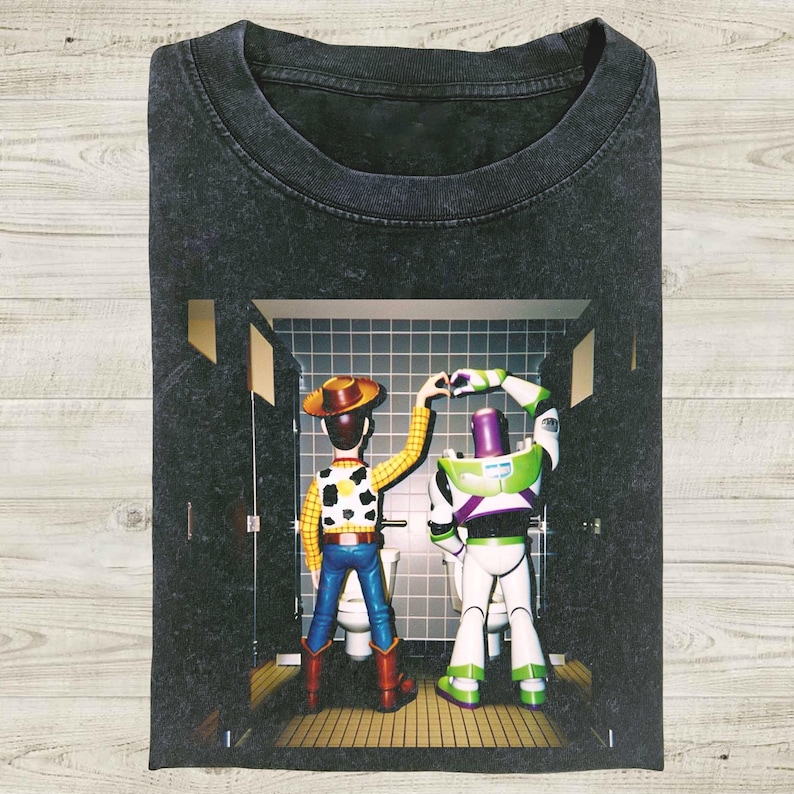 K&ouml;nnte beinhalten: Dunkelgraues T-Shirt mit einer Grafik von Woody und Buzz Lightyear aus Toy Story. Die Charaktere befinden sich in einer Badezimmerumgebung, Woody tr&auml;gt einen Cowboyhut und Buzz seinen Raumanzug. Das Bild ist auf einem weichen, l&auml;ssigen T-Shirt gedruckt.