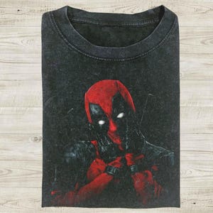 May include: A black acid-wash t-shirt featuring a graphic print of the character Deadpool. The character is depicted in a red and black suit with white eyes, shown from the chest up with hands positioned near the face in a pose.
