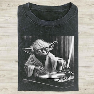 Op de afbeelding: Donkergrijs t-shirt met een zwart-wit afbeelding van Yoda als DJ, met een draaitafel en een lichtzwaard. De afbeelding toont Yoda in een gewaad, geconcentreerd op de platenspeler. Het shirt is opgevouwen en heeft een vintage wassing.