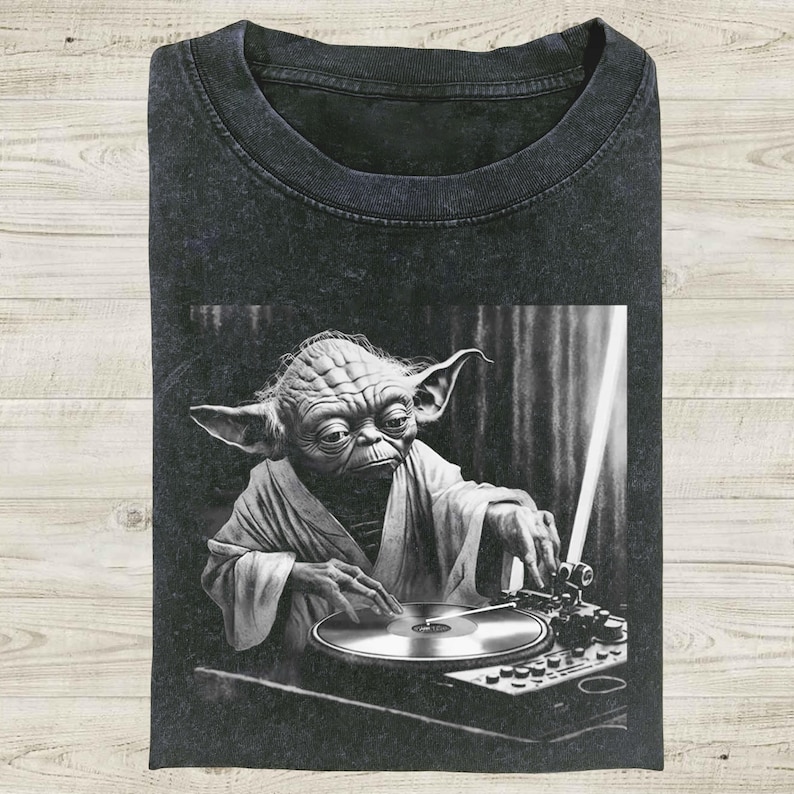 Op de afbeelding: Donkergrijs t-shirt met een zwart-wit afbeelding van Yoda als DJ, met een draaitafel en een lichtzwaard. De afbeelding toont Yoda in een gewaad, geconcentreerd op de platenspeler. Het shirt is opgevouwen en heeft een vintage wassing.