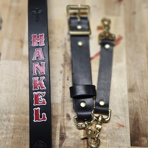May include: Black leather suspender straps with gold-colored hardware. One strap features the name "HANKEL" in red lettering. The suspenders have a classic design with a buckle and a clip for attachment, suitable for various outfits.