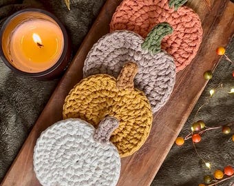 May include: A collection of five handmade crochet pumpkin coasters in various fall colors: orange, gray, yellow, and white. Each coaster features a stem and is arranged on a wooden serving board. A lit candle and decorative berries complete the autumn-themed display.