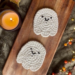 Puede incluir: Dos posavasos de fantasma de ganchillo hechos a mano, de color blanquecino, con caras bordadas en negro. Los posavasos están sobre una tabla de cortar de madera, con una vela encendida en un recipiente marrón oscuro en el fondo. El tema general es Halloween.