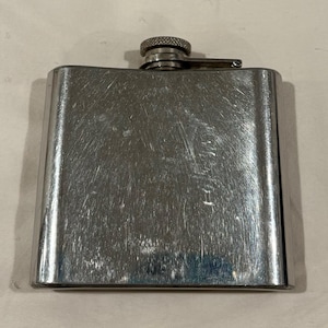 Puede incluir: Una petaca rectangular de acero inoxidable con tapón texturizado y cierre de bisagra. La petaca tiene un acabado de metal cepillado con arañazos visibles. El tapón es de metal texturizado más oscuro.