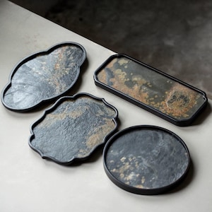 以下が含まれることがあります： 雲、長方形、円形など、さまざまな形状の4つの装飾用サービングトレイ。トレイは、黒い縁取りのある暗く質感のある表面を持っています。表面は、グレー、ブラウン、ゴールドの色合いでまだら模様になっています。