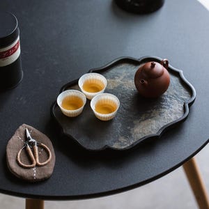 Puede incluir: Naturaleza muerta de un juego de té sobre una mesa negra. El juego incluye una pequeña tetera marrón, tres tazas de té y una bandeja decorativa. Unas tijeras pequeñas descansan sobre un posavasos marrón. También se ve una lata de té negra.