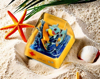 May include: A clear resin block with a yellow border, depicting two blue and yellow cartoon characters riding a surfboard on a blue wave. The block is sitting on a sandy surface with a starfish and seashell.