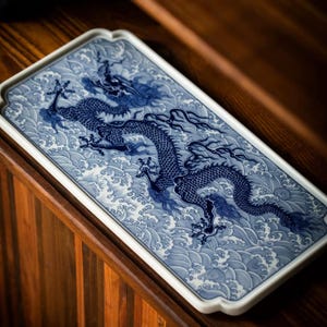 Op de afbeelding: Een rechthoekige keramische schaal met een blauw-wit drakenontwerp. De draak is gedetailleerd afgebeeld tegen een achtergrond van gestileerde golven. De schaal heeft een witte rand en staat op een houten oppervlak.