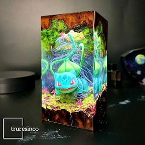 May include: A green and blue Pokemon character, known as Venusaur, is encased in clear resin with a brown wood base. The resin is filled with green and blue swirls and a few red mushrooms.