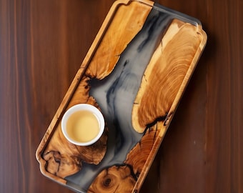 Puede incluir: Una bandeja de servir de madera con una incrustación de resina epoxi gris y negra. La bandeja tiene un acabado de madera natural y un borde redondeado. Una pequeña taza blanca con un líquido amarillo claro está en la bandeja.