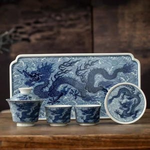 Puede incluir: Un juego de té de porcelana azul y blanca con diseño de dragón. El conjunto incluye una tetera con tapa, dos tazas, un posavasos y una bandeja rectangular. El dragón está representado con detalle sobre un fondo de olas.