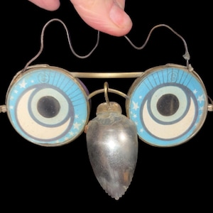 May include: A pair of novelty glasses featuring round frames with blue and white crescent moon and star graphics on the lenses. A metallic, teardrop-shaped nose piece hangs from the center bridge, and a thin wire frame connects the two circular lenses.