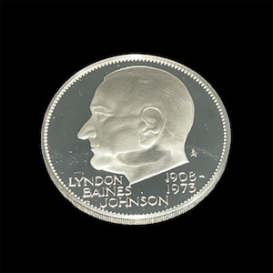May include: A silver coin featuring a profile of Lyndon Baines Johnson, with the text "LYNDON BAINES JOHNSON" and the years "1908-1973" around the edge. The coin has a detailed, textured border and is set against a black background.
