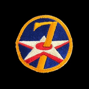 May include: A circular embroidered patch featuring a blue background with a yellow border. Centered is a white five-pointed star with a red circular center, overlaid by a large yellow number seven that extends from the top to the bottom edge.