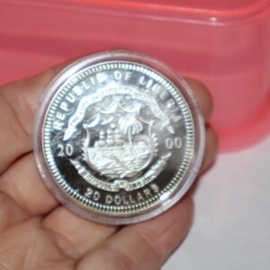 May include: A silver coin with the inscription "Republic of Liberia" and "20 Dollars" around the edge. The coin features a ship sailing on the water with palm trees in the background.