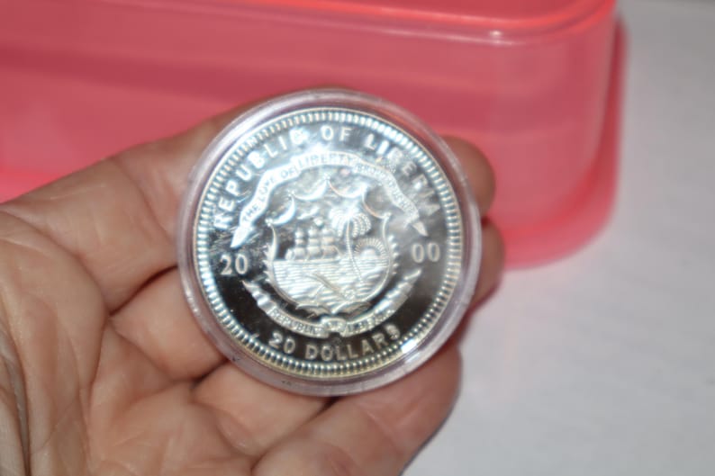 May include: A silver coin with the inscription "Republic of Liberia" and "20 Dollars" around the edge. The coin features a ship sailing on the water with palm trees in the background.