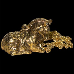May include: A gold-toned metal pendant shaped like a resting zebra with detailed engraved stripes across its body. The pendant features a small circular loop at the top for attachment and is connected to a matching gold-toned metal chain link.