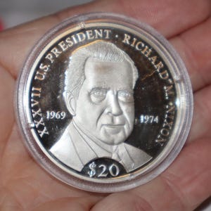 May include: Silver commemorative coin featuring a portrait of Richard Nixon, the 37th US President. The coin has the text "XXVII US PRESIDENT RICHARD M. NIXON 1969 1974" around the edge and "$20" in the center.