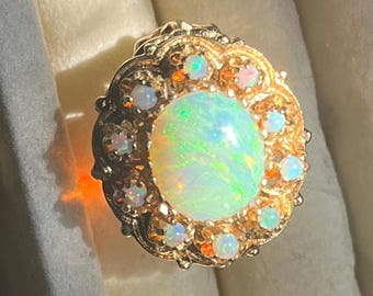 May include: A gold ring featuring a large, oval opal gemstone at the center, surrounded by smaller opals. The central opal displays a vibrant play of color, with hints of green and blue. The ring has an ornate, filigree design.